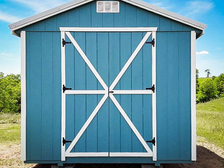 Blue utility shed with white trim and barn-style double doors, shown in a grassy field under a blue sky.