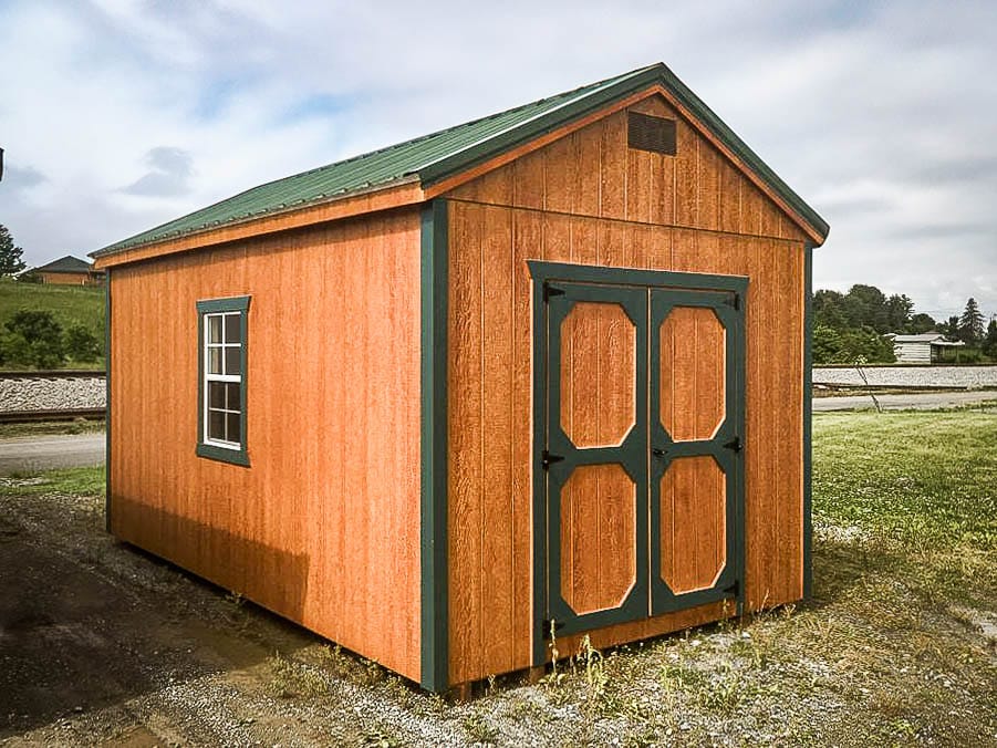 Wood-toned shed with green trim, gable roof, and double doors, placed on a gravel lot near open land.