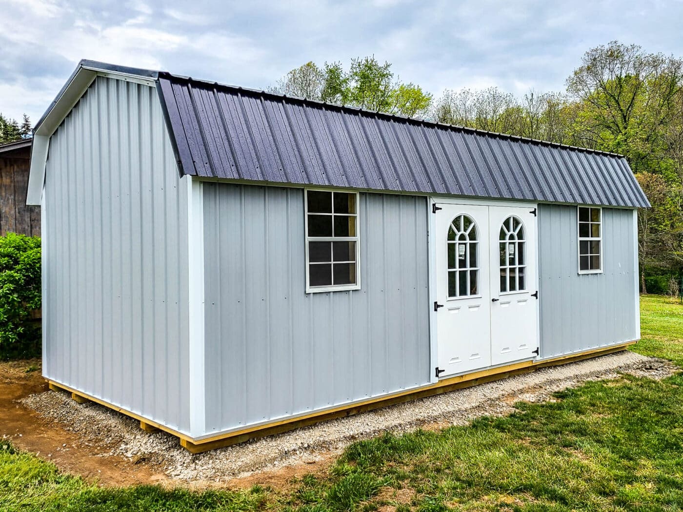 Gray lofted barn shed with white doors, arched windows, and black metal roof on a gravel pad.