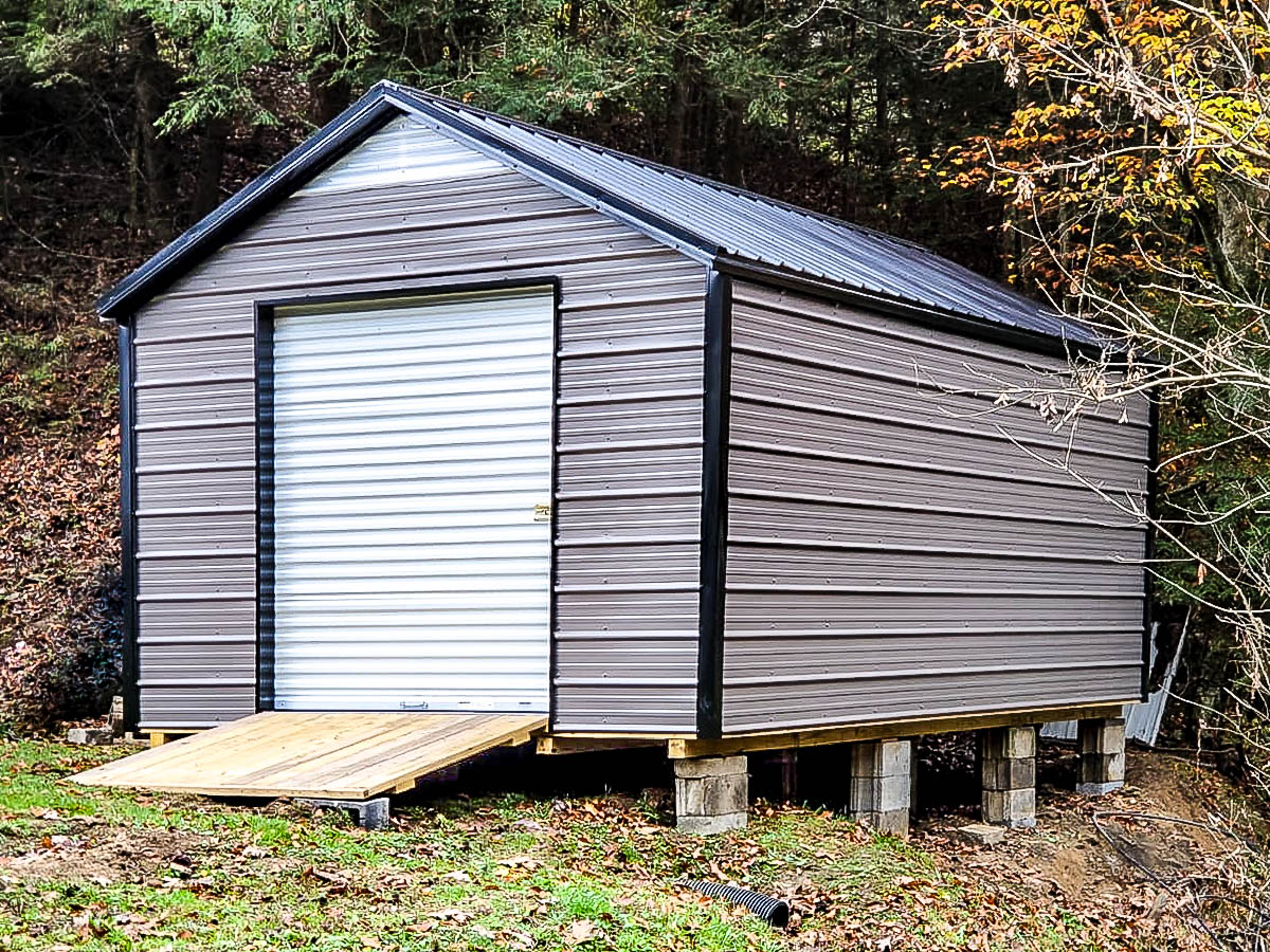 Elevated metal shed with gray siding, black trim, and a roll-up white door, with a wooden ramp for accessibility.