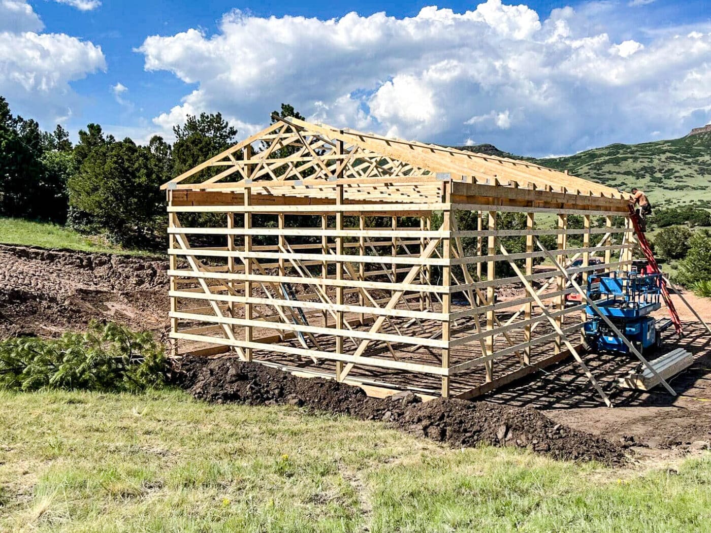 A partially built wooden pole barn framing with an exposed frame and roof trusses, situated in a rural setting with rolling hills and trees in the background. A blue tractor is parked beside the structure, and the construction site has visible dirt and grass patches.