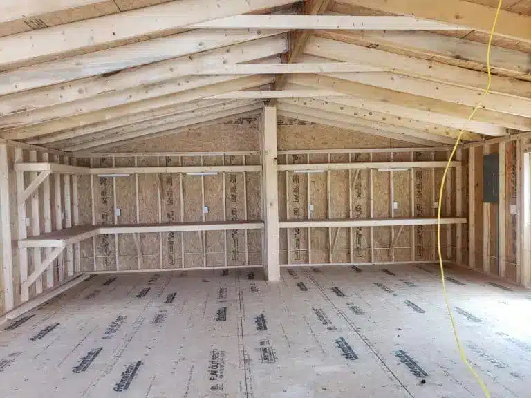 An unfinished interior of a wooden pole barn with exposed framing, rafters, and wall studs. The ceiling is supported by wooden trusses, and the floor is covered with protective sheathing material. Horizontal wooden beams run along the walls, possibly for future siding or shelving installation. The space is open and well-ventilated, allowing for various customization options.