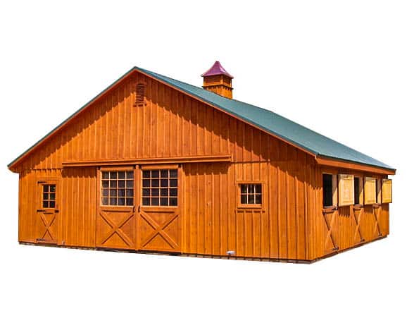 A fully constructed wooden pole barn with a warm-toned natural wood exterior, a green metal roof, and a decorative cupola. The barn features large sliding doors with an X-brace design, multiple windows, and a covered entry area.