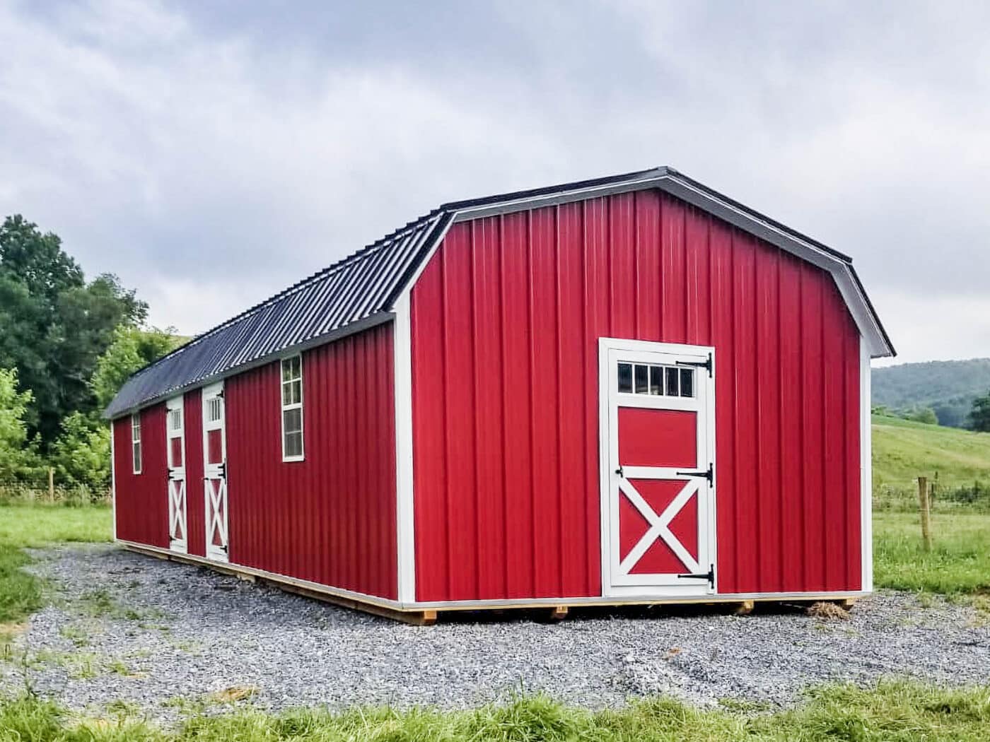 Red barn-style shed with white X-brace doors, vertical metal siding, and black metal roof on a gravel base.