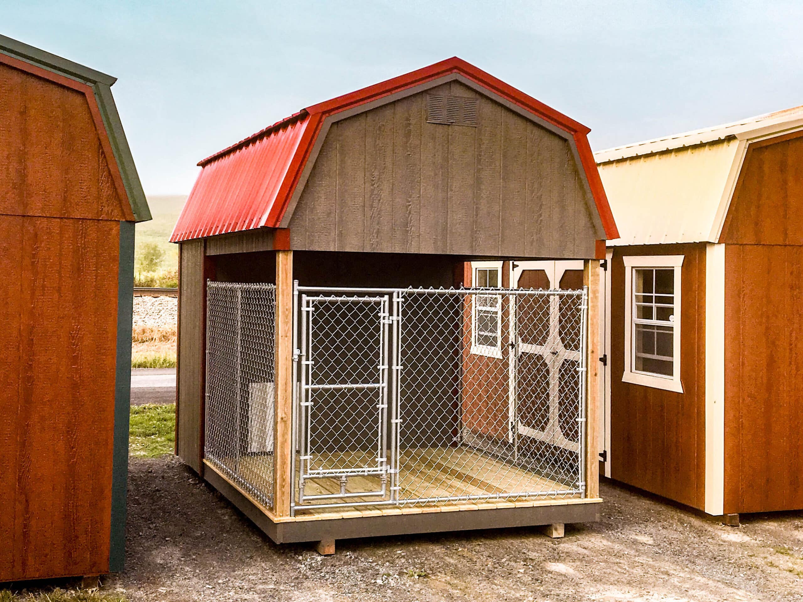 A small outdoor dog kennel with a red roof, chain-link fencing, and a sturdy wooden base, situated between two larger storage sheds.