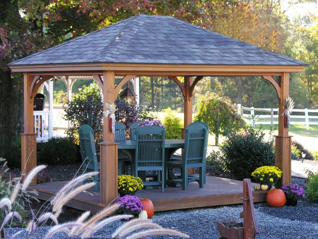 Brown wooden Pavilion with dark gray roof over green table and six matching chairs - orange pumpkins yellow and purple floral bunches at base - lush woodsy area beyond white fence in background