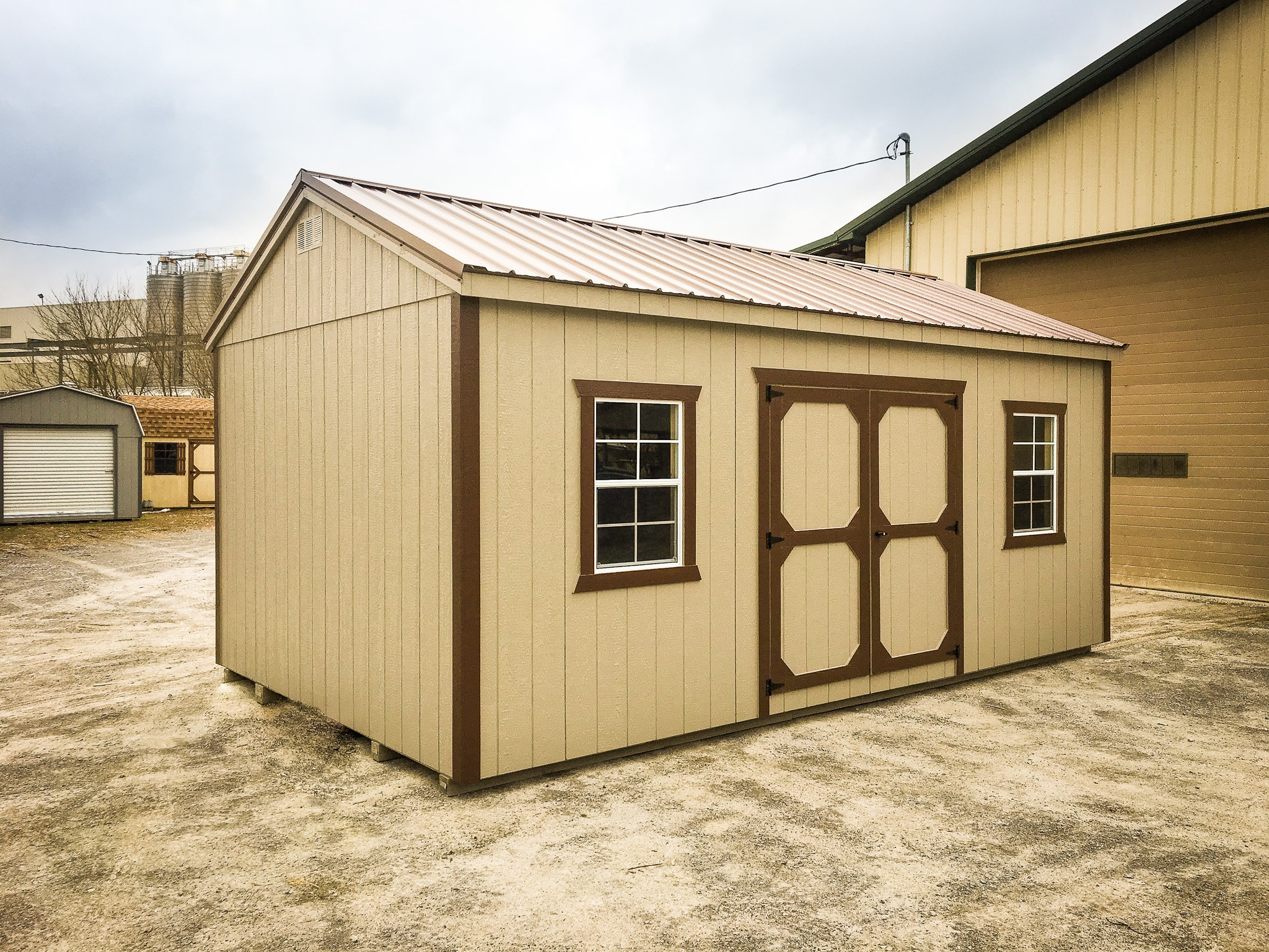 Beige shed with brown trimmed double doors and windows under brown metal gable-style roof on lot with other brown and gray sheds in background