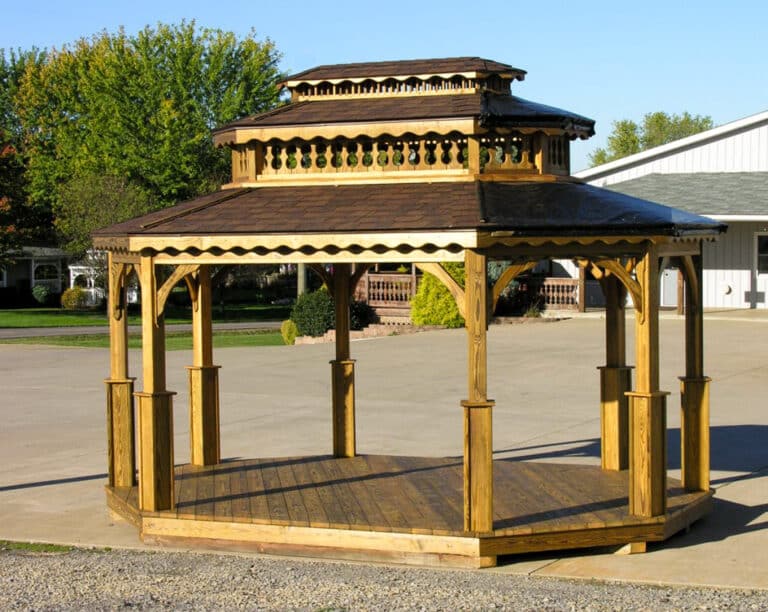 One of our luxurious wooden pavilions for sale in Roanoke VA - Eight wooden beams extend from platform to support triple-tiered roof with scalloped wooden trim - set on concrete pavement with patches of grass and a few trees in the distance