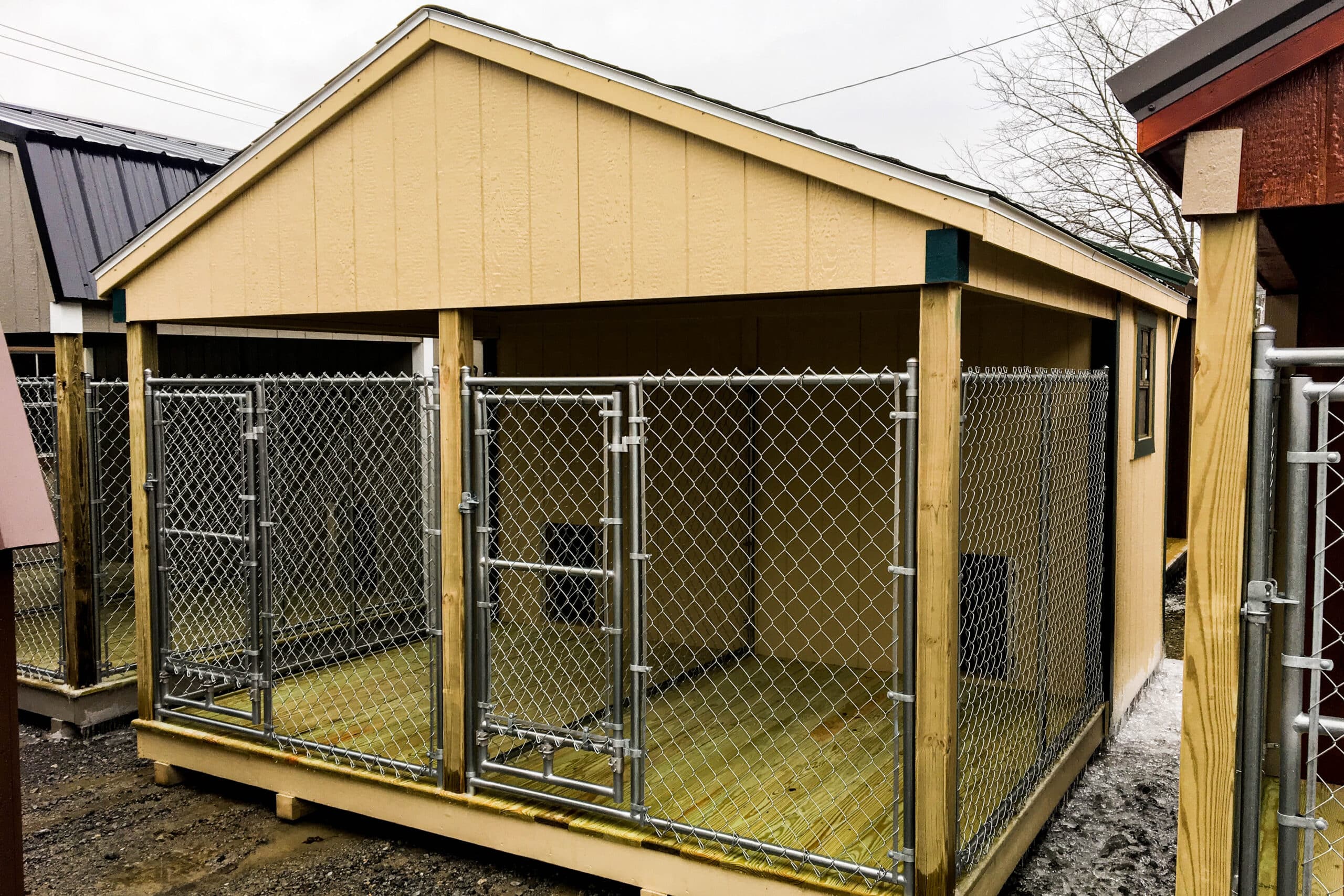 A large wooden dog kennel with a light yellow exterior and a sloped roof. The structure includes two separate outdoor sections enclosed by sturdy chain-link fencing with individual gates. The interior flooring appears to be made of wooden planks, providing a clean and elevated surface. The kennel is situated outdoors, with a partially overcast sky and neighboring structures visible in the background.