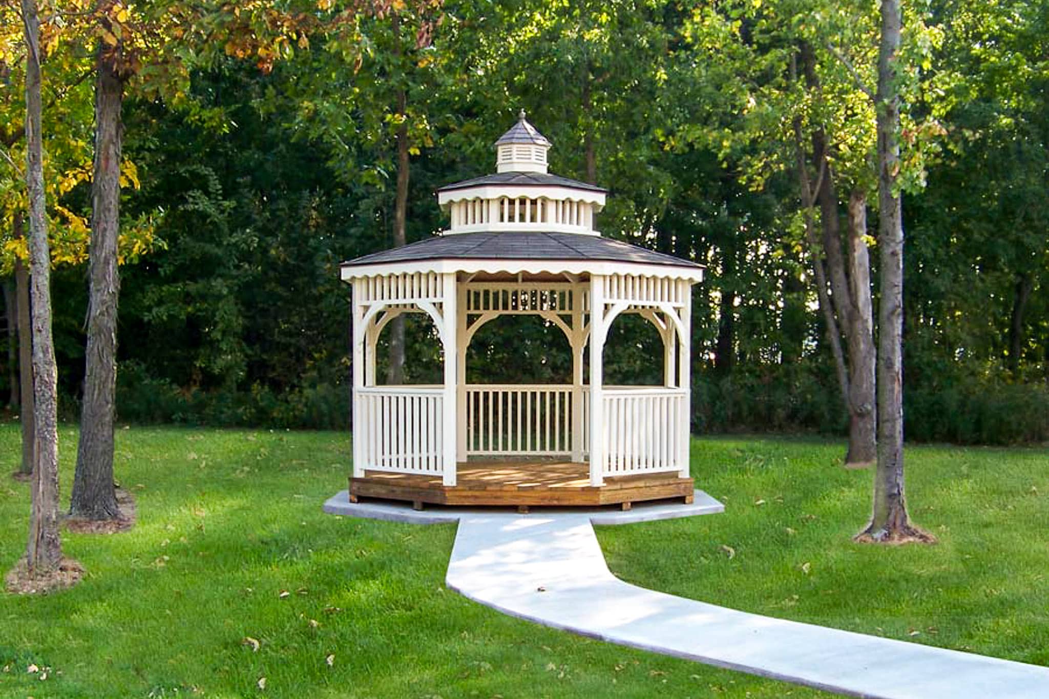 white gazebo with tho-tiered gray shingled roof with cupola on top - gazebo sits at the end of a short light gray concrete path on grass with trees in background